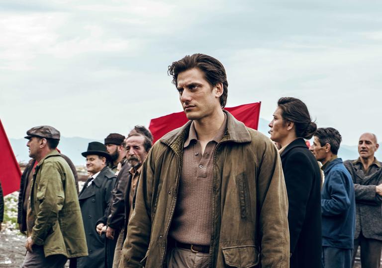 Luca Marinelli stands in muted clothes in front of people and red flags in a field in the film Martin Eden