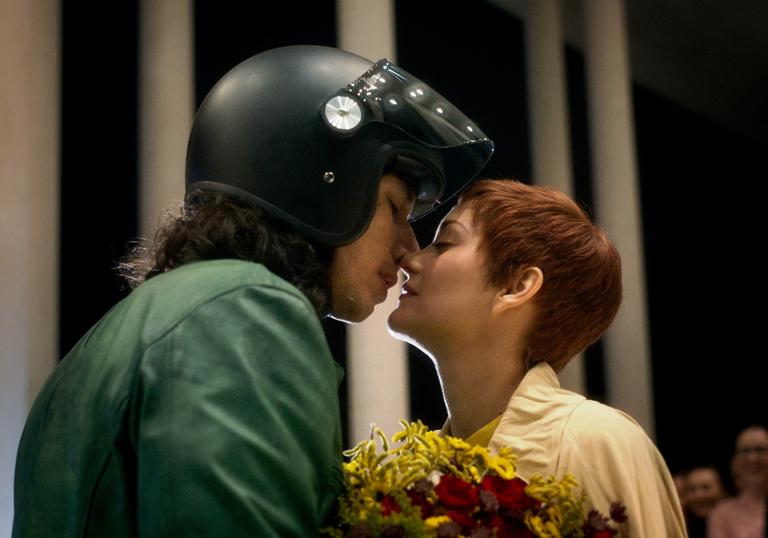 Adam Driver wears a black motorcycle helmet with his nose to Marion Cotillard, who is holding a bunch of flowers, in Leos Carax's Annette