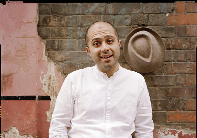 Mahan Esfahani smiling in front of a wall, with his hat hanging on the wall behind him