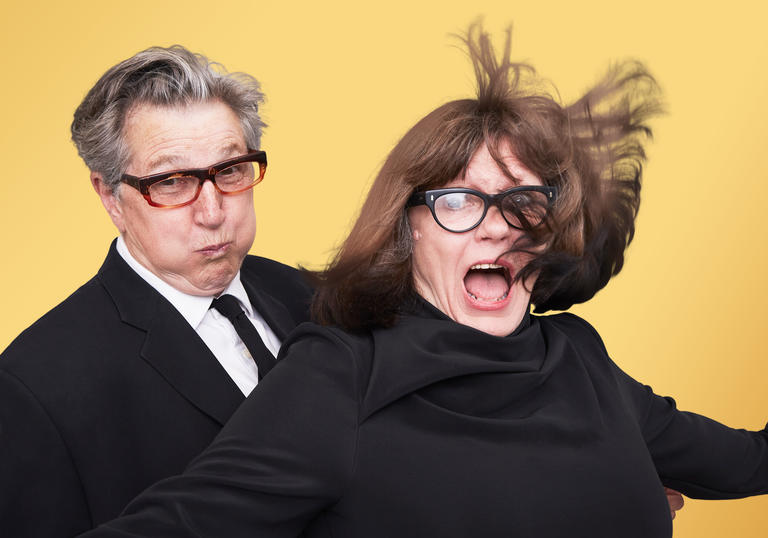 Two women stand next to each other. One is wearing a suit blowing air into their mouth with their lips pursed. She is wearing a black suit and tie. The other lady is wearing a black top and has her mouth wide open and her short bob brown hair is blowing in a gust of wind.