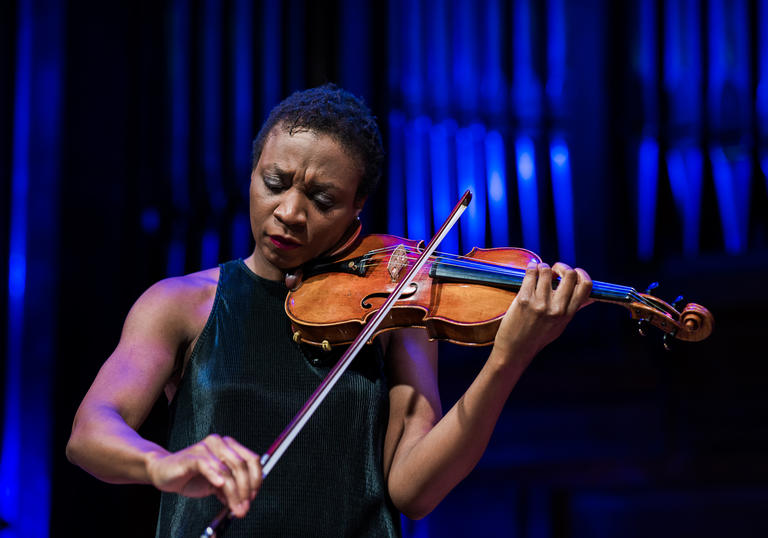 Tai Murray stands playing the violin, in front of organ pipes with a blue light on them