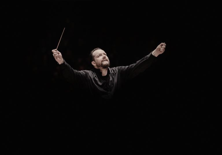 Andris Nelsons with his arms raised. He's holding a conductor's baton in his right arm, and he appears to be coming out of the black background.