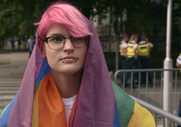 Tobi, a young person with pink hair, is draped in the rainbow flag in front of a few police officers in a still from Colors of Tobi