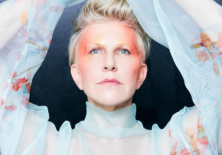 Joyce DiDonato in a blue floaty top with her arms above her head, with red face paint on her temple and the sides of her face