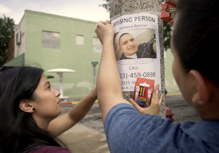 A still from Missing in Brooks County, two people putting up a missing poster on a lamppost