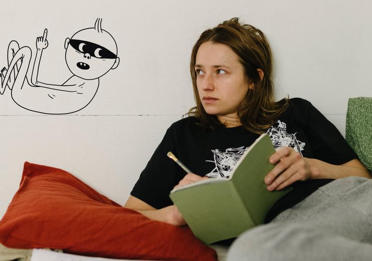 A young woman writes in a notebook while looking at an animated baby on a plain wall, in a still from Ninjababy