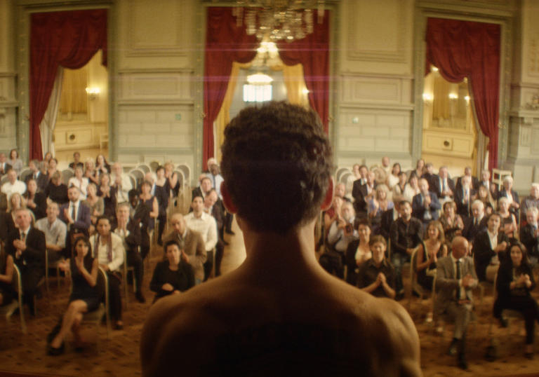 A still from The Man Who Sold His Skin, a man with a naked torso stands facing a large audience in a grand room