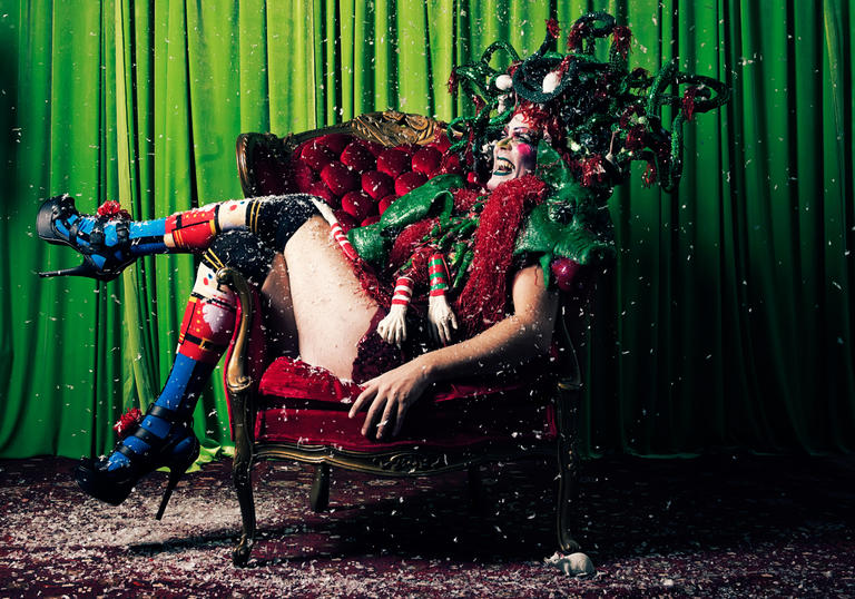Taylor Mac sitting on a red chair behind green curtains wearing a Christmas-style head piece