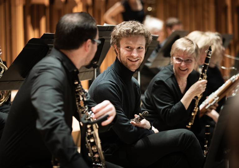 Two members of Britten Sinfonia smile at another member, who has his back to the camera. They are all on the Barbican stage, holding their instruments.