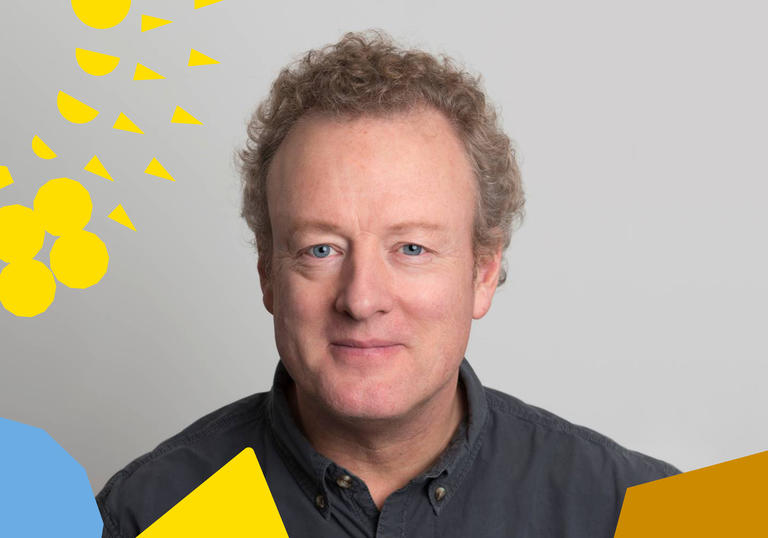 Howard Goodall smiling on a white background, with colourful shapes around the edge of the photo (BBC brainding)