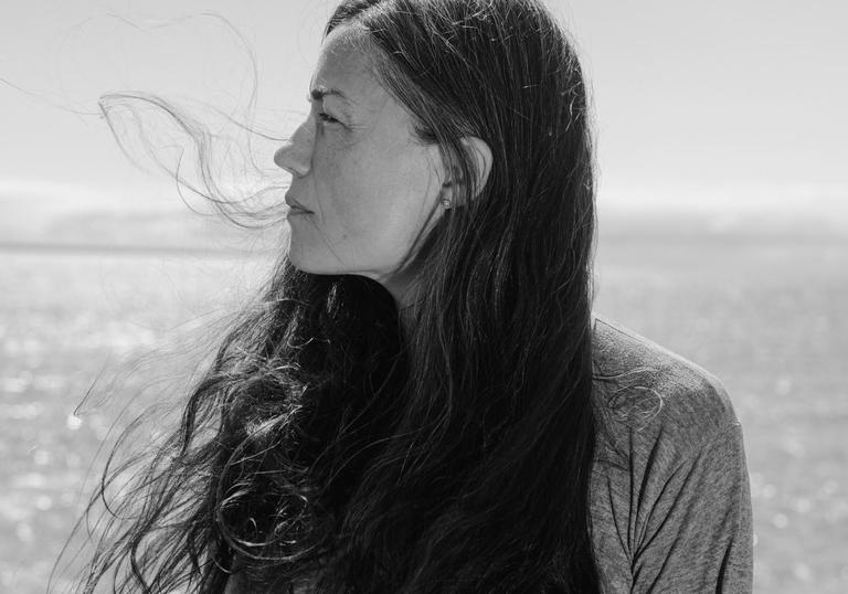 A black and white portrait photo of Grouper. The sea is behind her, her long hair is blowing in the wind and she is looking of her left shoulder.