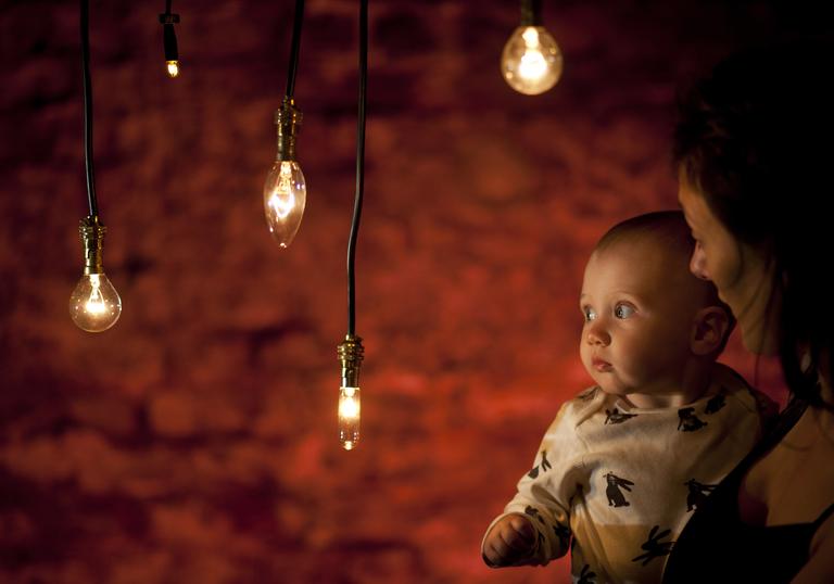 A baby in the arms of their mother look at hanging tungsten style light bulbs, bathed in a warm glow. Behind them is a red exposed brick wall. The mother is looking intently at the child.