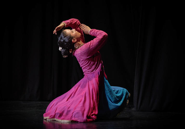 A dancer dressed in pink, kneels on a blacked out stage