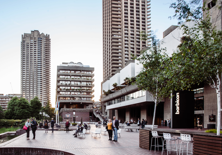 A photo of the barbican centre by the lake, and the tower block building in the background with groups of people standing.