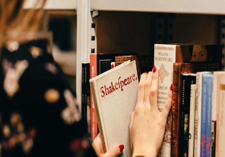 A person is taking a white book from a shelf. The book says 'Shakespeare' on the front in red text.