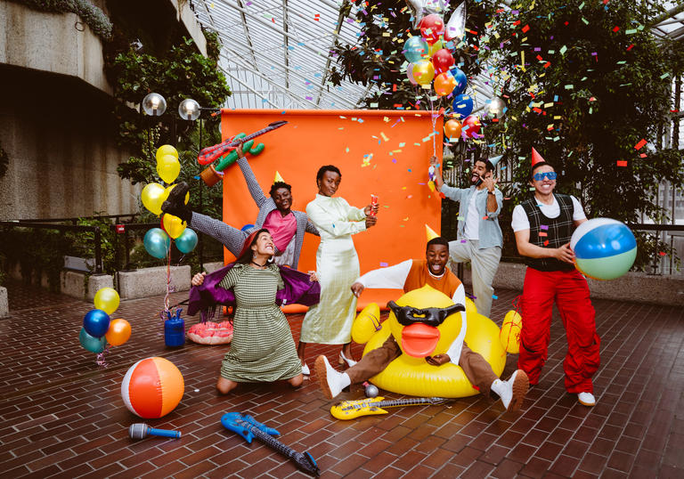 A party of people in front of an orange screen wearing party hats, dancing with confetti and inflatables.