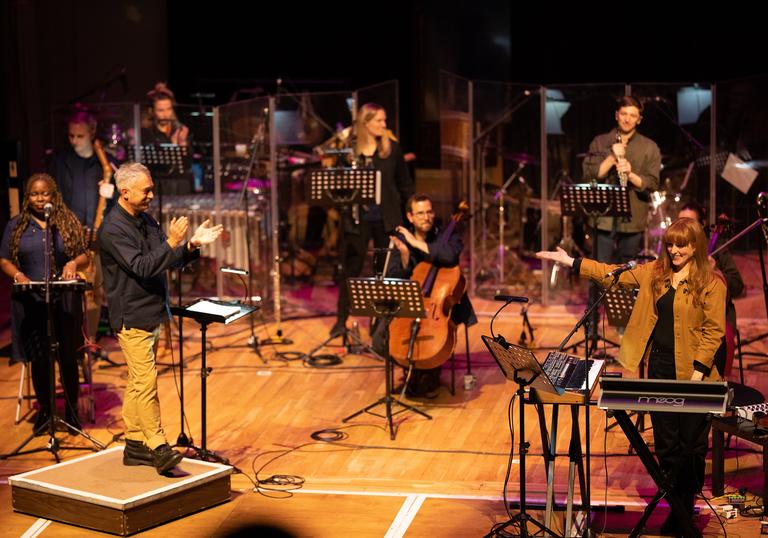 Hannah Peel performing with Paraorchestra. From behind a keyboard on the left of the stage she is gesturing to the ensemble who are applauding.