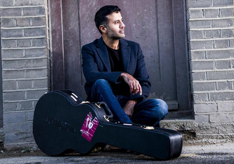 Hamza Namira sitting in a doorway with his guitar case on the floor in front of him.