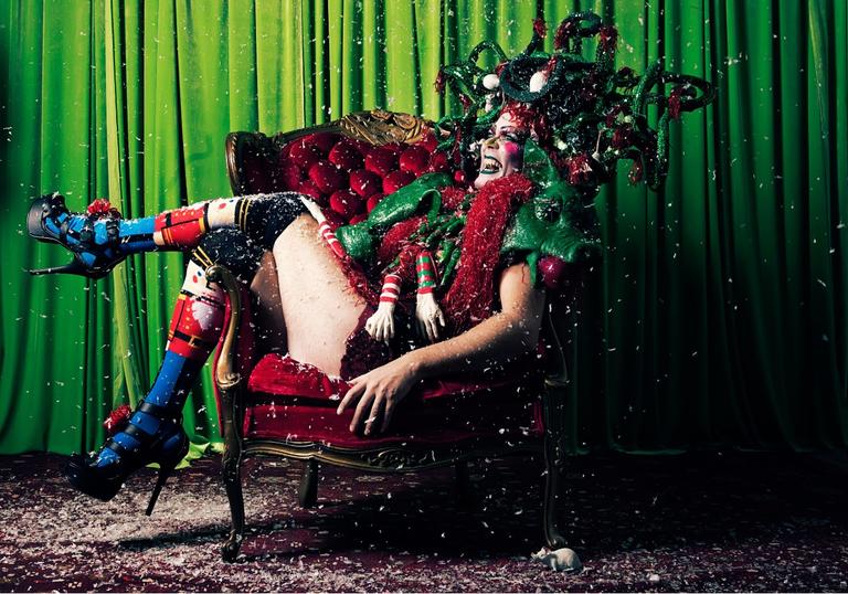 a colour photo of Taylor Mac in festive drag reclining over an armchair against a green backdrop