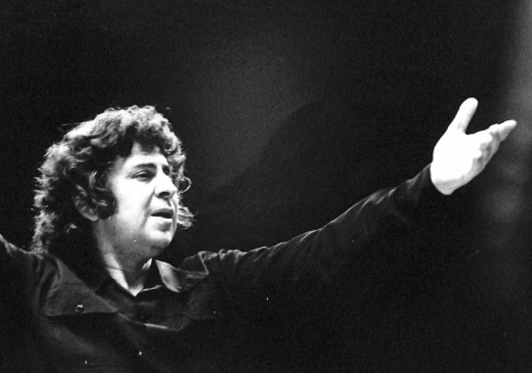 Mikis Theodorakis with arms outstretched conducting. He is wearing a black shirt