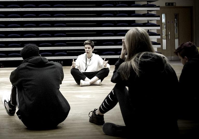 A black and white image of a theatre workshop with an instructor facing participants