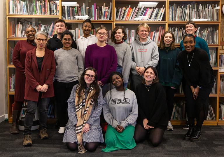 A group of people stand in a row in front of a bookshelf with three people kneeling on the floor in front, they are all facing forward to the camera smiling