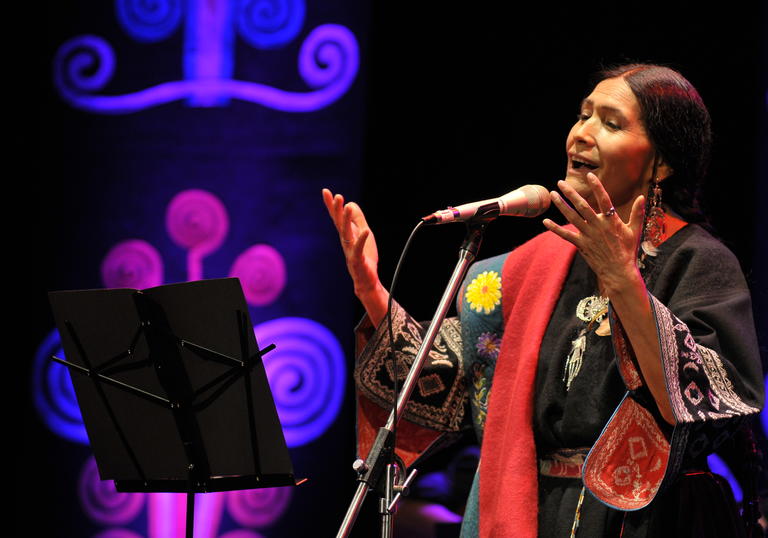 Luzmila Carpio singing, she is wearing a colourful traditional indigenous outfit with her hands outstretched in front of her