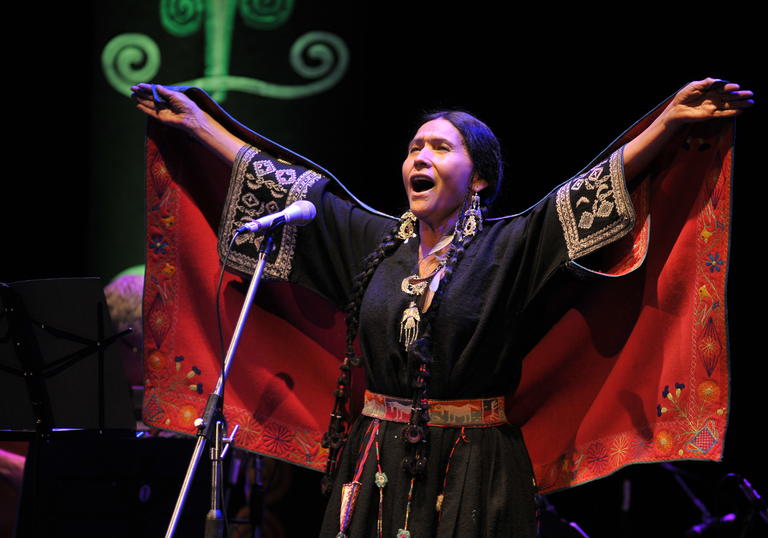 Luzmila Carpio singing, she is wearing a colourful traditional indigenous outfit with her hands outstretched above her