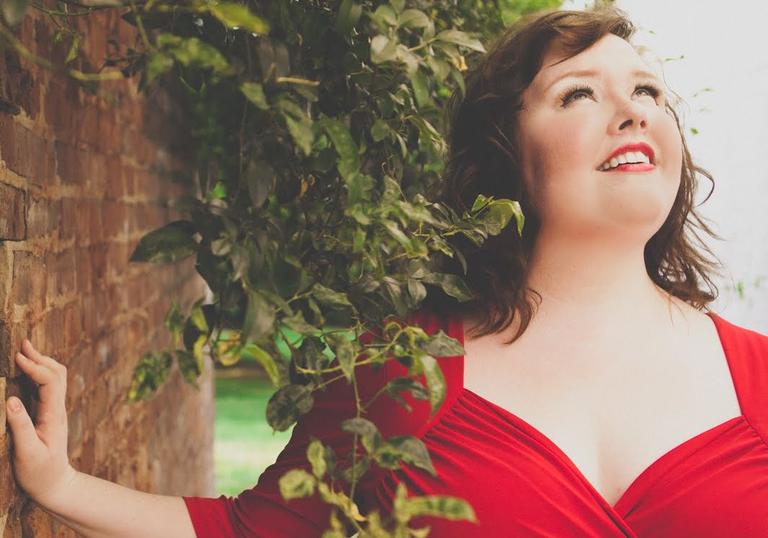 Jamie Barton in a red dress beside a brick wall and a plant draping over her shoulder