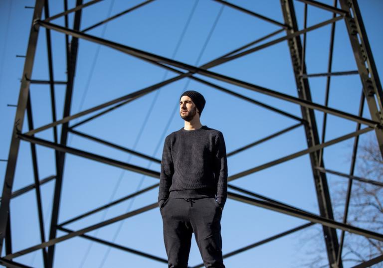 a colour photo of Manu Delago standing in front of an electricity pylon and a blue sky