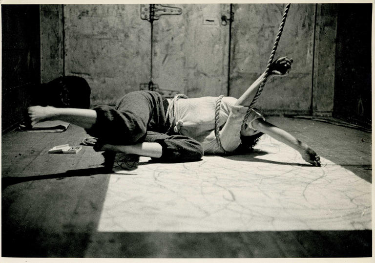 An image of Carolee lying on the floor with her arms tied up in some rope 