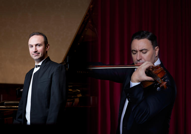 In the left of the image Simon Trpceski stands smiling in black tie in front of a piano. In the right of the image Maxim Vengerov is playing his violin.