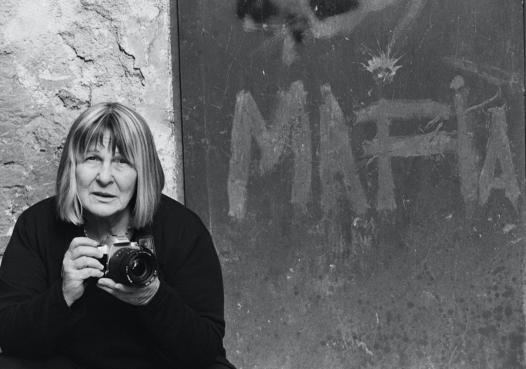 A black and white image with a person with blonde short hair and a fringe stands in front of an image that says Mafia