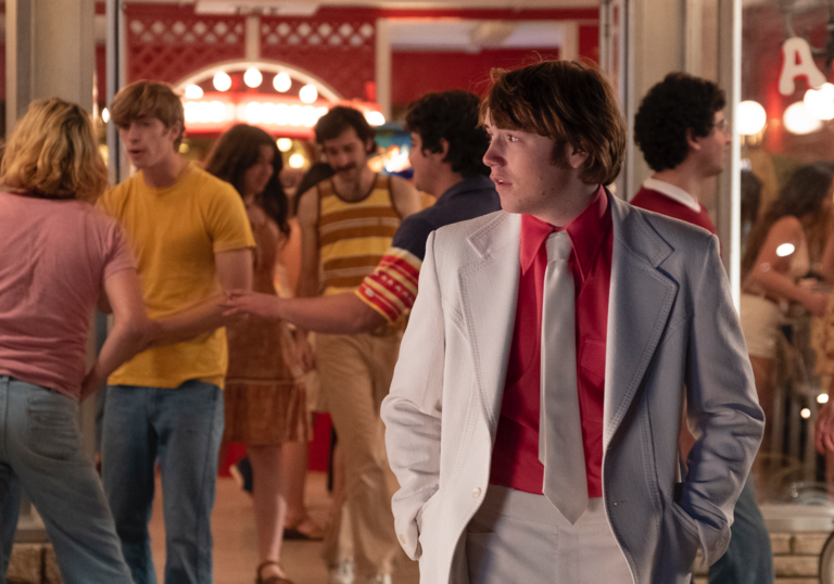 Cooper Hoffman in his role as Gary, standing outside a busy theatre wearing a white suit