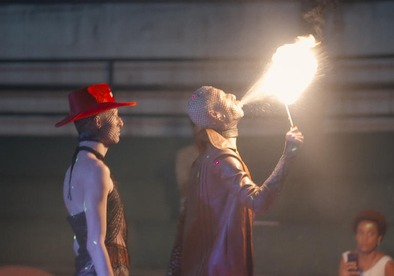 Two young queer people wearing mesh, one blowing fire