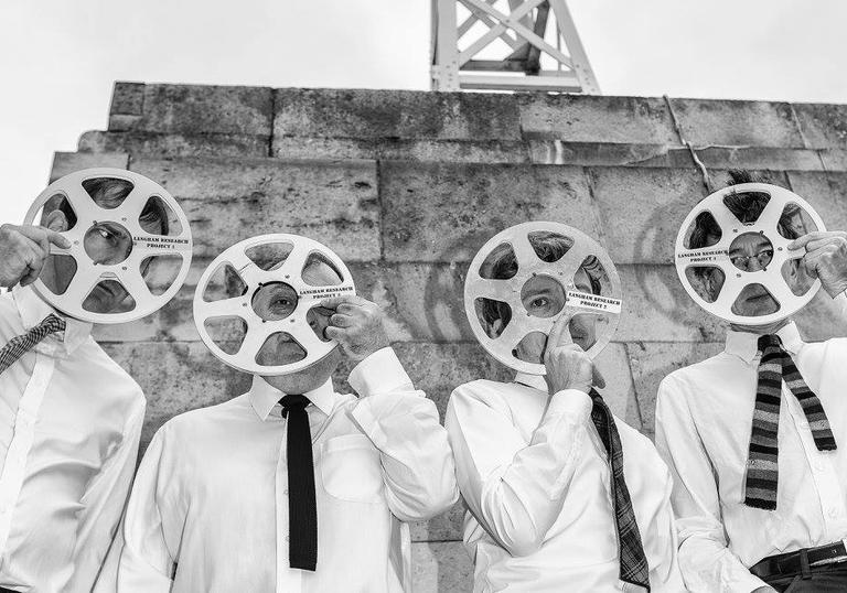 A black and white image of four men dressed in shirts and ties, holding tape reels over their faces