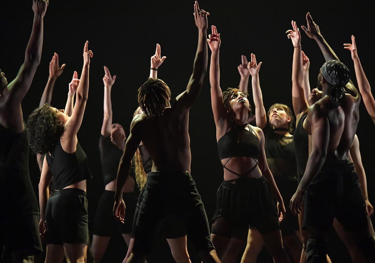 Dancers dressed in black stand together with their hands raised in the hair.