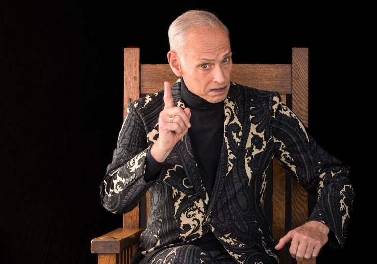 A photo of John Waters sitting on a chair and wagging his finger