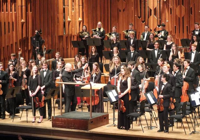 The London School Symphony Orchestra standing on stage in the Barbican Hall holding their instruments