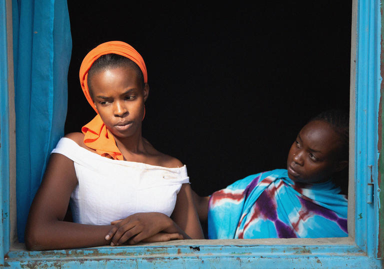 Framed by a blue open window sill, a daughter looks out longingly while her mother attempts to comfort her