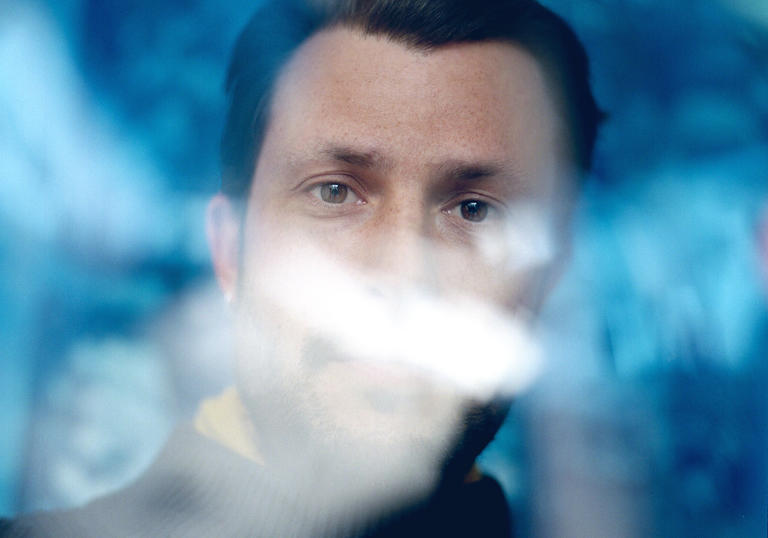 Orlando Weeks in front of a blue background with some distortion across his face. He is wearing a black turtleneck jumper.