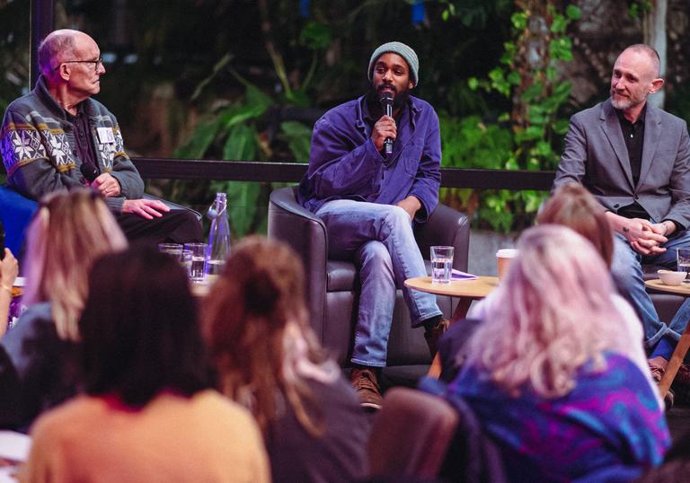 A person sits in the middle of two other people on a stage with a microphone in their hand, they are talking directly to a group of people sat in front of them