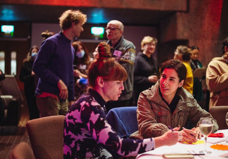 Two people sit around a table talking to each other, with their arms on the table. Two people stand behind them having a conversation