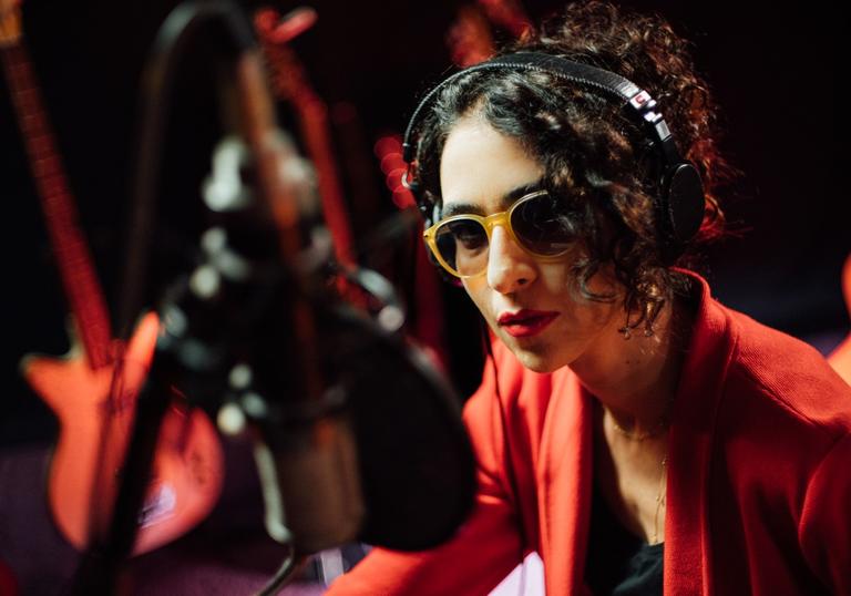 a close up photo of Marisa Monte wearing sunglasses and a red suit against a dark background