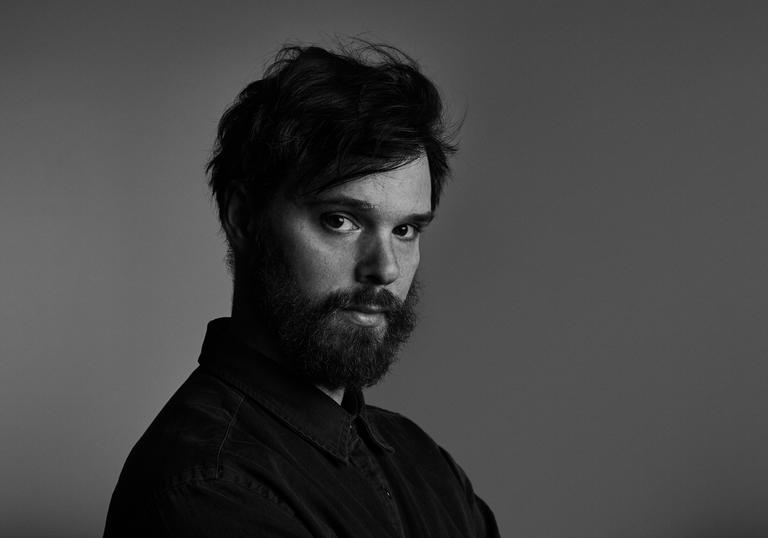 A black and white portrait of Dave Longstreth, looking sideways at the camera. He is wearing a dark shirt and has a beard.