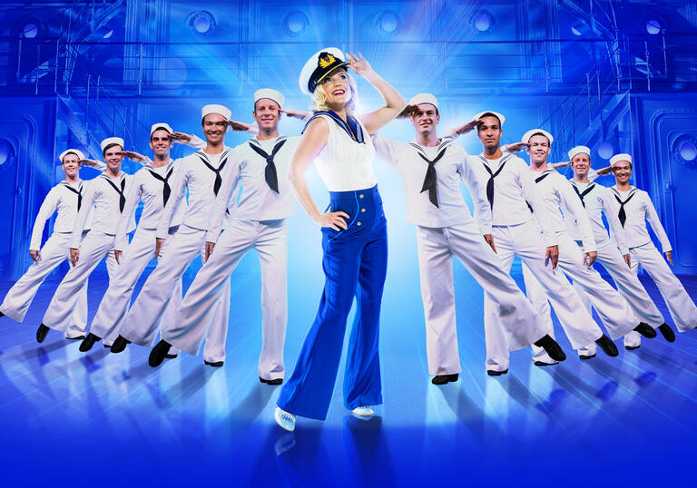 Kerry Ellis dressed in a nautical themed outfit and hat as Reno Sweeney. She stands surrounded by sailors, all saluting. 