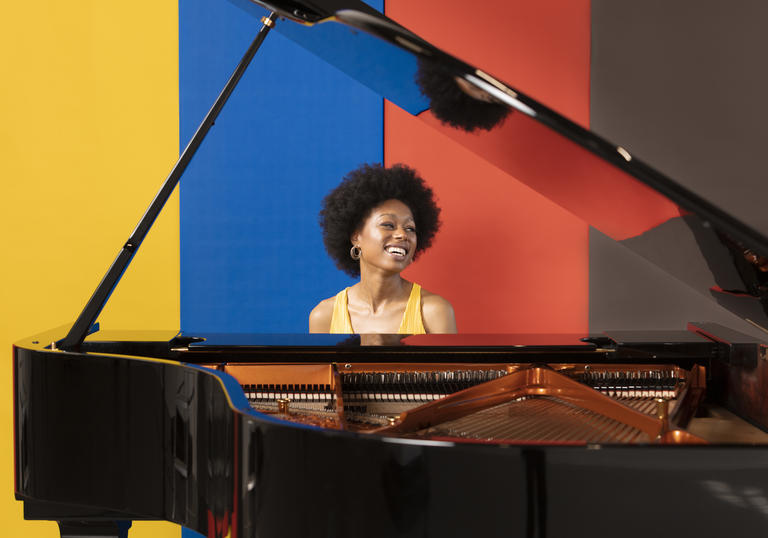 Isata Kanneh-Mason sits at a piano, smiling and looking left. Behind her is a backdrop of yellow, blue, red and black stripes. 