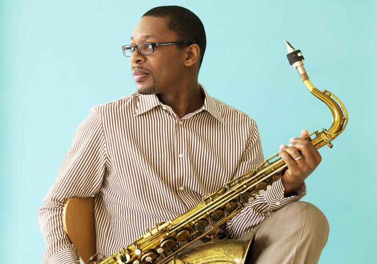 Ravi Coltrane sitting crosslegged holding his saxophone and looking over his shoulder