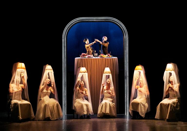 Six performers in white dresses sit on chairs under shaded lamps while two figures appear to be eating dinner together above them at a table. 