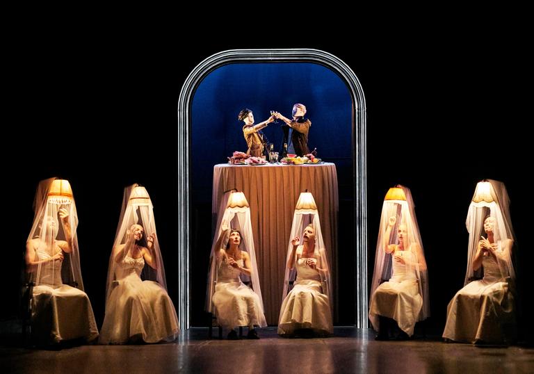 Six performers in white dresses sit on chairs under shaded lamps while two figures appear to be eating dinner together above them at a table.